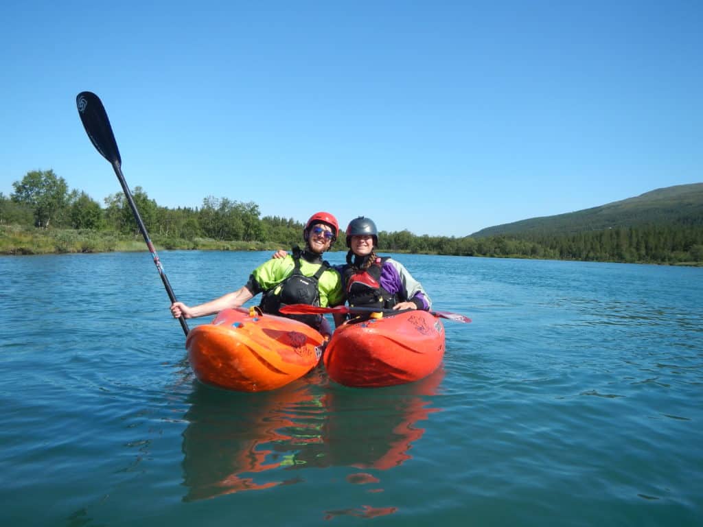 A man and a woman in matching kayaks on flat water in Norway. It is very sunny and they are smiling.