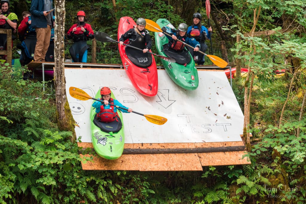 Three female kayakers are falling down a ramp located above a river. A woman in a green kayak is in front with two other women in red and green kayaks behind her.