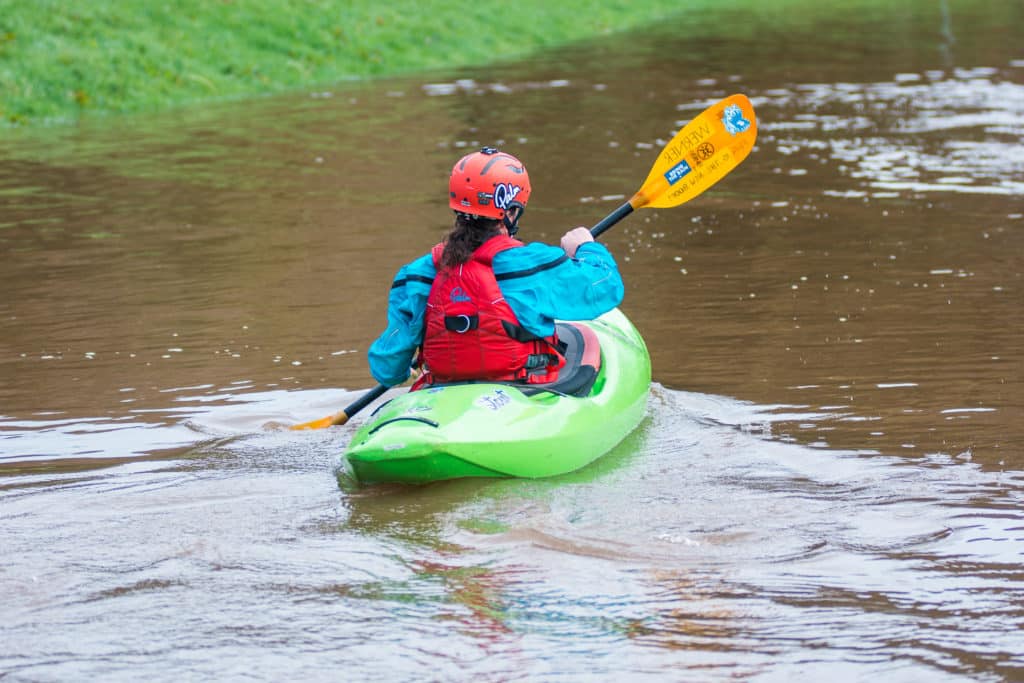 A girl paddles in a green Waka kayak on a flat lake.