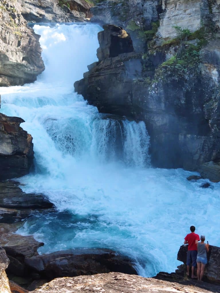 An impressive waterfall. At the bottom, two people stand facing it. They are small in comparison.