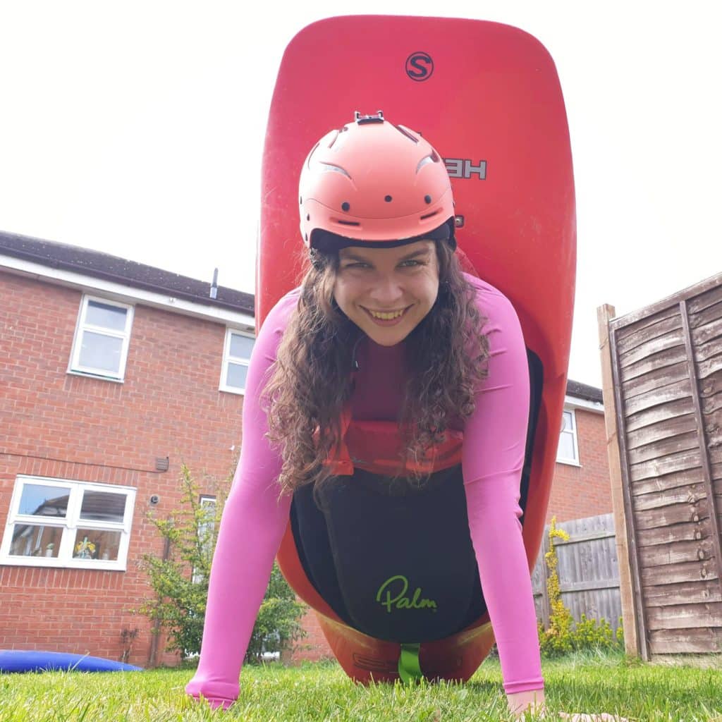 A girl is sitting in a red playboat in her garden. She has pushed the boat onto its end and is supporting herself with her arms in a press up position.