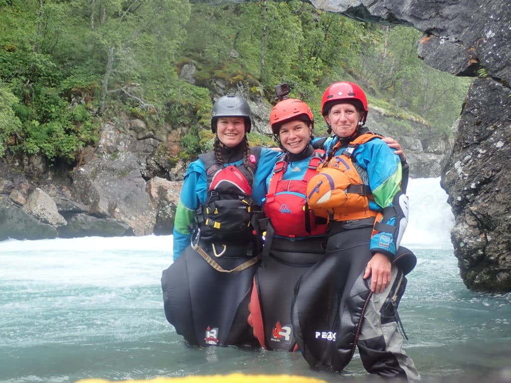 Three females kayakers stand below a drop. They are knee deep in the water and smiling.