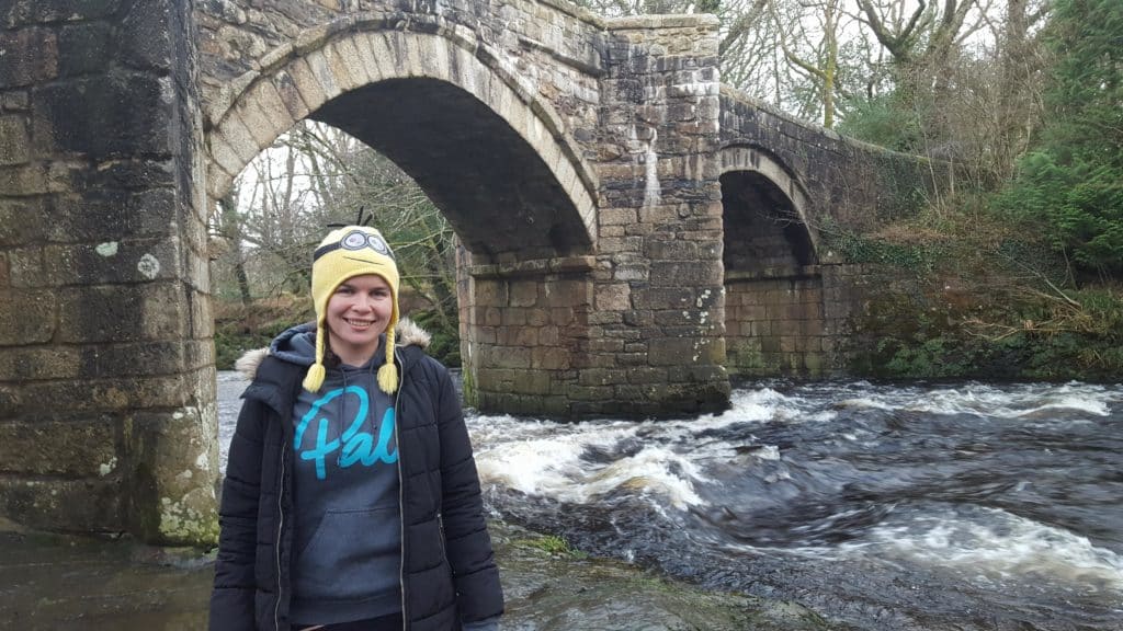 Girl standing by a bridge with whitewater underneath it. The girl is wearing a yellow minion hat and a hoody that says Palm.