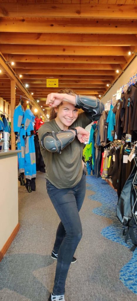 A woman standing in a kayaking shop. She is wearing elbow pads and is standing in a warrior pose.